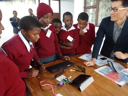 株式会社ict4e 代表 原 秀一 氏 （右端）、2016年にタンザニア連合共和国アルーシャ地区のさくら女子中学校で実施されたプログラミング教育の一コマ、中央にある基盤がむき出しのデバイスは、福井で開発された子供向けパソコン「 IchigoJam 」