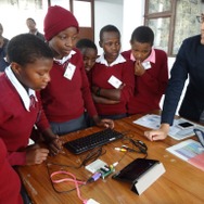 株式会社ict4e 代表 原 秀一 氏 （右端）、2016年にタンザニア連合共和国アルーシャ地区のさくら女子中学校で実施されたプログラミング教育の一コマ、中央にある基盤がむき出しのデバイスは、福井で開発された子供向けパソコン「 IchigoJam 」
